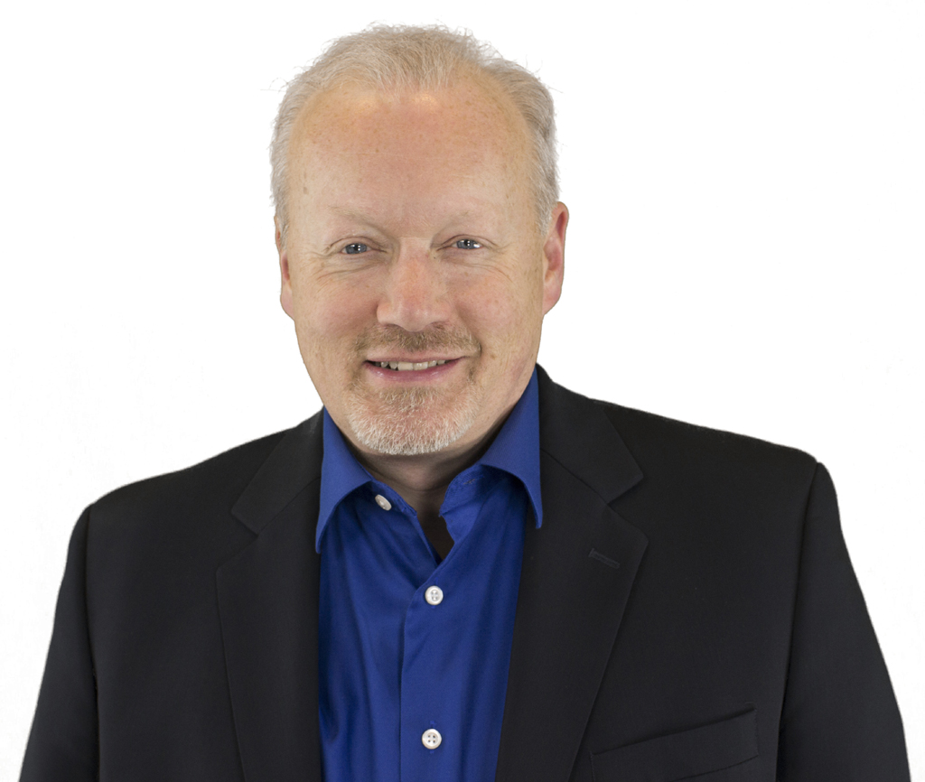 Mike gives a friendly smile in a headshot. He is a white man with blue eyes, gray hair and a beard. He wears a black blazer over a blue collared shirt. 